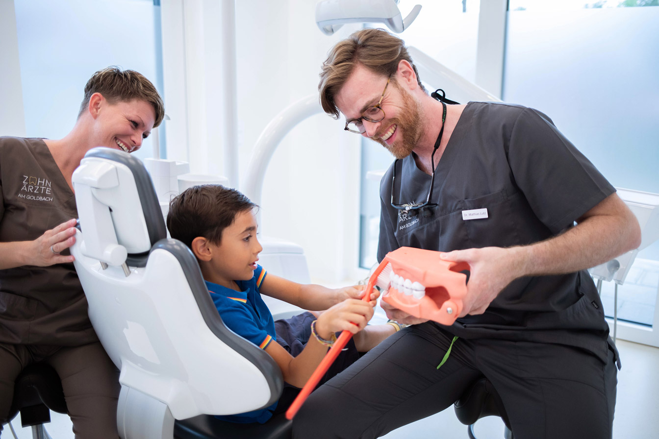 Zahnärzte am Goldbach, Dr. Lutz zeigt einem Jungen an einem Gebiss wie man richtig Zähne putzt